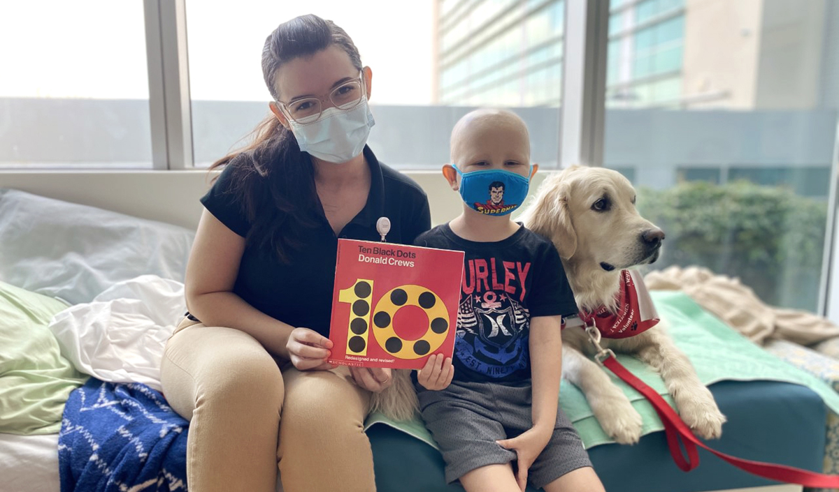 UCF PedsAcademy intern sitting with child patient and therapy dog on hospital bed