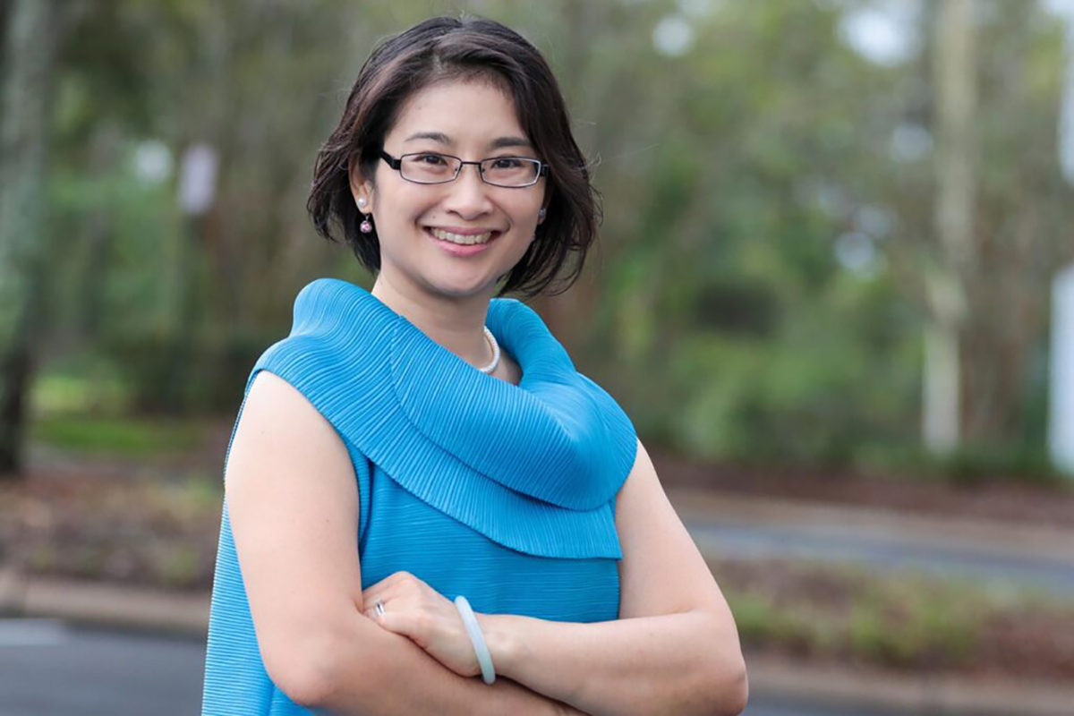 Su-I Hou wearing a blue blouse and smiling against a blurred nature background.