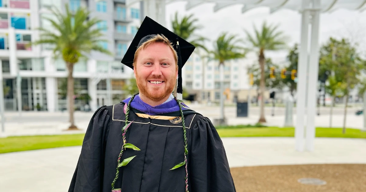 Carter Gresham, wearing graduation regalia, smiles in front of a backdrop of buildings and palm trees.