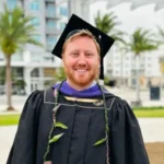 Carter Gresham, wearing graduation regalia, smiles in front of a backdrop of buildings and palm trees.