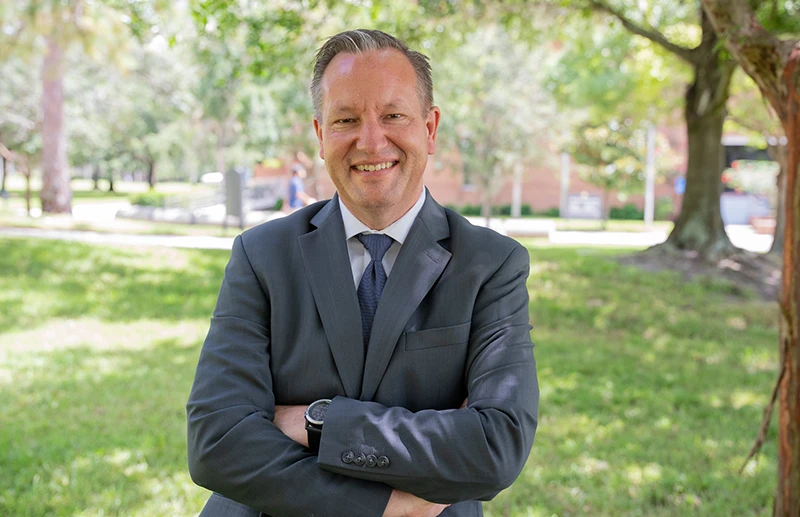 Dr. Bradley Heim, wearing a dark gray suit, stands smiling in front of greenery outside with crossed arms.
