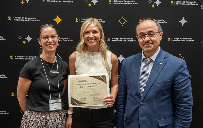 Kristina Childs, Mackenzie Masters and Naim Kapucu standing in front of a black CCIE-branded backdrop. Masters is holding a Research Impact Showcase presenter certificate.