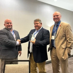 Three men dressed in sports jackets stand smiling behind chairs in a conference room. Professor Jeremy Hall is in the middle, holding an awards plaque and shaking hands with the man to his right.