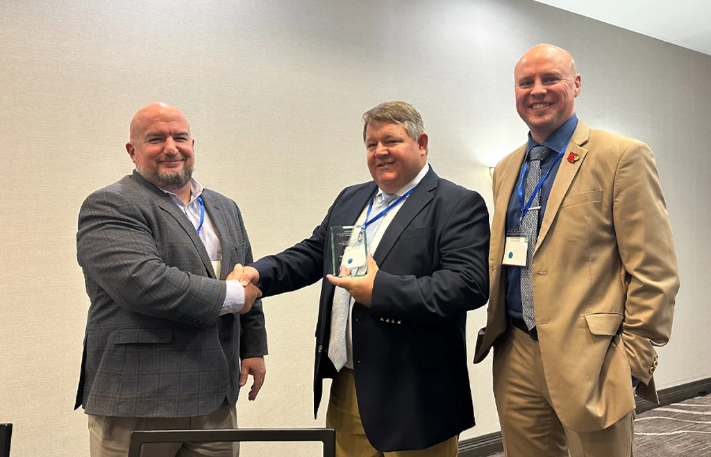 Professor Jeremy Hall, center, accepts the Donato J. Pugliese Award from members of the Southeastern Conference for Public Administration’s awards committee. (Courtesy photo) Three men dressed in sports jackets stand smiling behind chairs in a conference room. Professor Jeremy Hall is in the middle, holding an awards plaque and shaking hands with the man to his right.