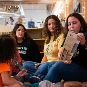 Three women from UCF’s School of Teacher Education sit on a classroom floor reading a book to young children listening in front of them.